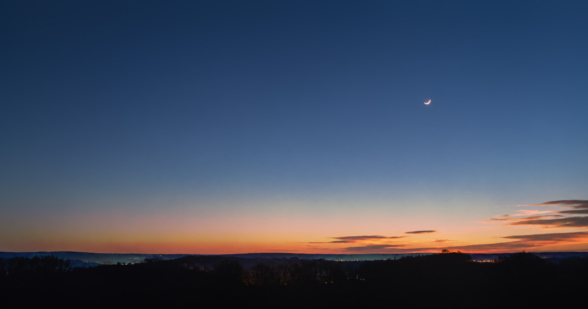 A thin new moon barely visible in a dark, quiet sky over a soft horizon